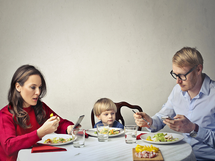 Mother and father using technology at dinner table, while ignoring thier young child with them