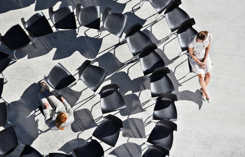 Two business people sit apart amongst a spiral of empty chairs.
