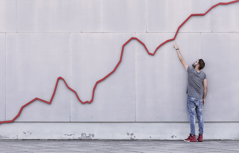 A red bar line graph rises like a financial graph or graphic with a young man pointing at the growth against a wall