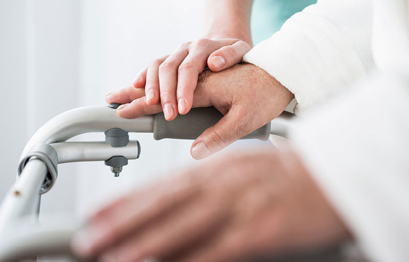 La main d'un soignant est posée sur celle d'un aîné utilisant une marchette.